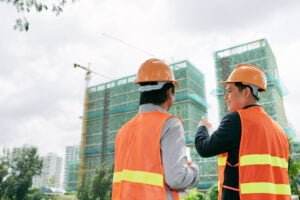 Two construction workers in orange safety vests and hard hats stand outdoors, facing partially constructed high-rise buildings with cranes. One worker points towards the buildings, teaching safety measures as they discuss progress, while the other listens attentively. Trees and cityscape are visible.