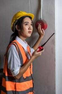 A woman wearing a yellow hard hat and an orange safety vest is holding a two-way radio in one hand and pressing a red emergency button on the wall with the other, triggering visual alarms. She appears focused and attentive.