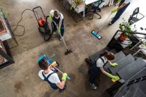 Aerial view of four people in uniform, equipped with cleaning tools and wearing green gloves, ensuring office safety by cleaning an office space with desks and plants. One person vacuums the floor, another mops, while two others wipe surfaces, including the stairs.