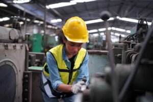 A worker wearing a yellow hard hat, a safety vest, and gloves is operating machinery in an industrial facility. The background features various industrial equipment and a spacious, well-lit interior with a metal roof, highlighting the importance of awareness regarding operation hazards.