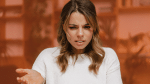 A woman with long brown hair in a white sweater stands indoors, looking confused. Her eyebrows are furrowed and her left hand is slightly raised, palm up, as if questioning something related to human resources. The background is blurred with warm, orange tones.