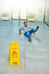 A man slips and falls on a wet floor in a hallway. He appears to be losing his balance while sitting on the floor, with one leg in the air. A yellow caution sign indicating "Wet Floor" is prominently placed in the foreground.