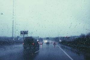 A rainy day view from inside a car, looking through the windshield covered with raindrops. Several vehicles with headlights on are visible on the road ahead. The overall scene is blurry due to the changing weather conditions.