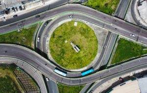 Aerial view of a roundabout with green grass in the center, intersected by multiple roads. Several vehicles, including cars and a bus, navigate the roundabout with highway driving safety tips and tricks in mind. Surrounding areas include buildings, paved roads, and greenery.