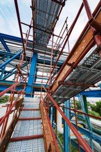A view looking up at a network of metal scaffolding and walkways at a construction site. The scaffolding, designed with safety in mind, consists of blue and orange support beams, with perforated metal planks forming the walkways. The background shows parts of a partially constructed building.