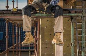 A construction worker in tan pants and tan boots sits on scaffolding, exemplifying the importance of working safely from heights. Their hands, wearing gloves, rest on their lap. The background features scaffolding, a wooden structure, and a ladder.
