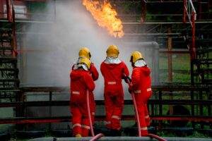 Three firefighters in red suits and yellow helmets stand side by side, using hoses to spray water on a fire. Flames are visible in the background, with a metal structure and some greenery around them. This scene reflects their rigorous training exercise or emergency response as part of the Three Sixty Safety program.