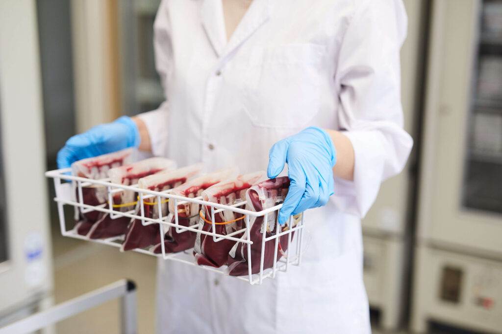 A person wearing a white lab coat and blue gloves is holding a white plastic basket containing multiple blood bags. The background appears to be a medical or laboratory setting with equipment and storage units, where strict protocols are followed to prevent bloodborne pathogens.