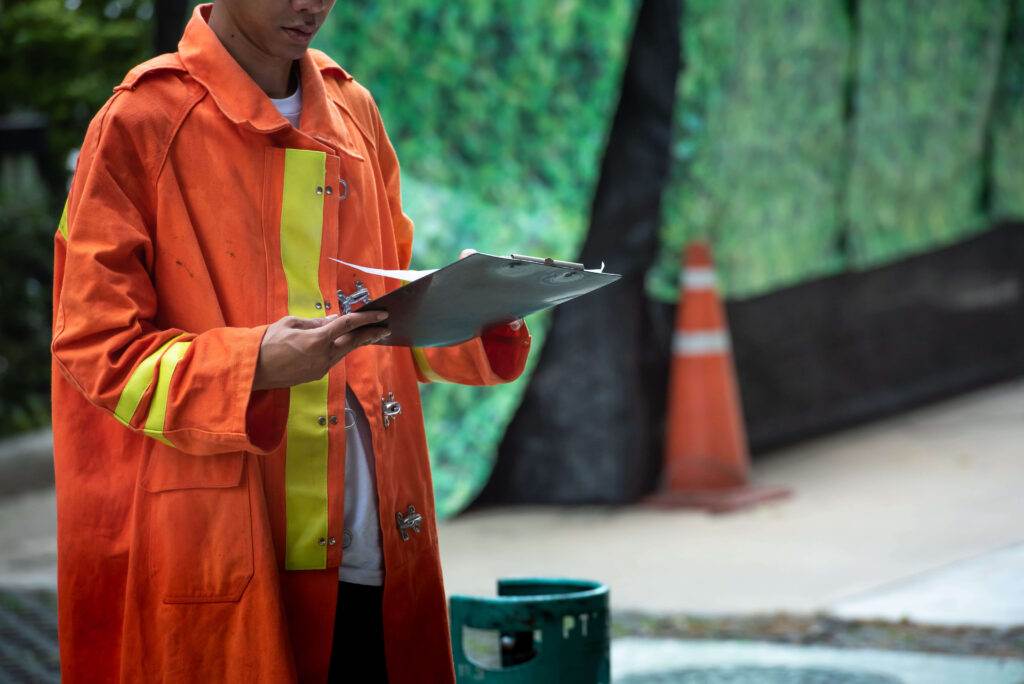 A person wearing an orange raincoat with yellow reflective stripes is holding a clipboard and writing on it. In the background, there is an orange traffic cone and a green barrier, suggesting that practice drills are underway at an outdoor work or construction site.