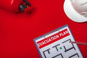 A red fire extinguisher and a white hard hat are placed on a bright red surface, ready for any emergency. A clipboard holds a paper labeled "EVACUATION PLAN," showing a basic floor plan with a pen pointing to a section of the map.