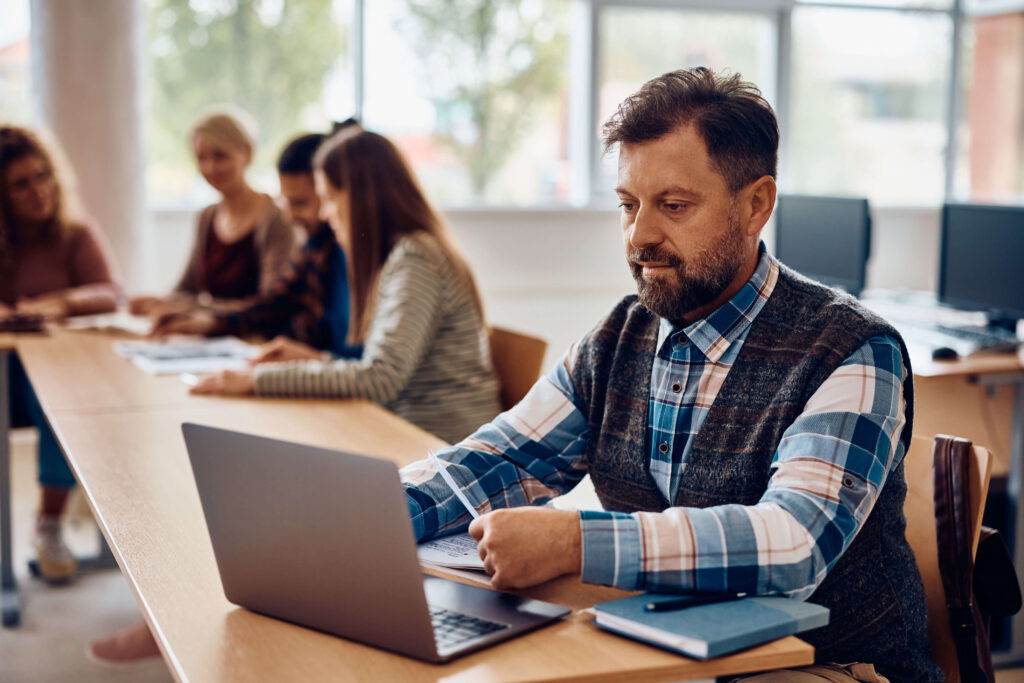 A man with a beard, wearing a plaid shirt and vest, works on a laptop at a long desk in a bright room. Several other people are seated along the desk in the background, engaged in e-learning discussions or working on papers.