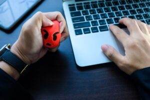 A person typing on a laptop with their right hand while squeezing a red stress ball with an angry face using their left hand, clearly illustrating the stress on the job. A smartphone is visible on the desk next to the laptop.