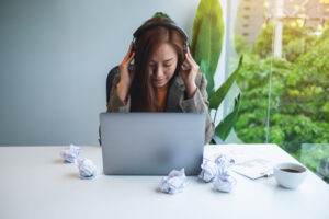 A person sits at a desk in front of a laptop, wearing headphones and looking downward, seemingly stressed from work-related pressure. Crumpled paper balls are scattered on the desk alongside a clipboard and pen. A cup of coffee and a large green plant add slight comfort to the tense atmosphere.
