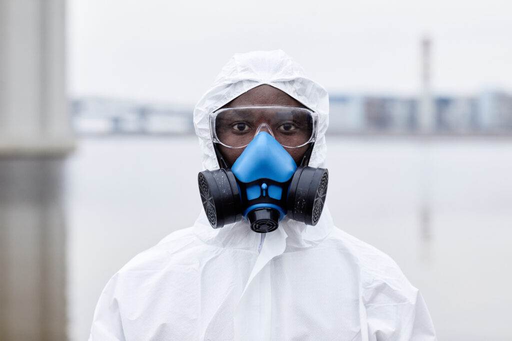 A person wearing a white hazmat suit, protective goggles, and a blue respirator mask stands outdoors near a body of water. The PPE is essential for mitigating biological risks. The background is slightly blurred, showing buildings and industrial structures in the distance.