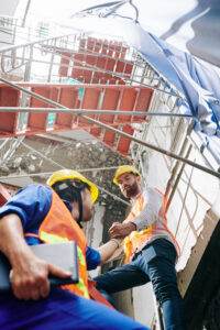 Two construction workers in safety vests and hard hats are in a building under construction. One worker helps the other climb a staircase made of scaffolding, ensuring fall protection. The background shows construction materials and debris.