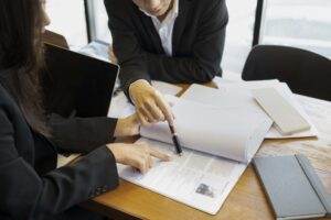 Two people in business attire are sitting at a wooden table, reviewing a document. One person is holding a book and pointing at the text with a pen, while the other person is also pointing at the document. A laptop, papers, and a notebook are on the table as they discuss important safety management protocols.