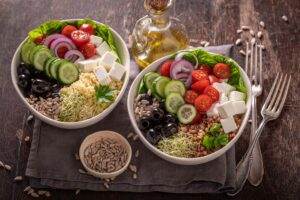 Two Mediterranean-style salad bowls filled with ingredients like quinoa, cucumber slices, cherry tomatoes, onion slices, feta cheese, black olives, and sprouts, garnished with parsley. An olive oil bottle and a small bowl of sunflower seeds sit nearby, promoting healthy eating. Utensils are at the ready.