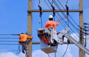 Two utility workers wearing orange uniforms and hard hats perform maintenance on power lines. One worker is in a bucket lift, using an insulated rod, while the other climbs a pole, employing fall prevention equipment to secure themselves. They exemplify safe work practices under a blue, cloud-studded sky.