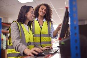 Two women wearing high-visibility safety vests are working together at a computer in an industrial setting. One is seated, typing on the keyboard, while the other stands and points at the monitor, smiling. Their teamwork enhances safety performance. Shelving and boxes are visible in the background.