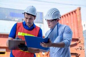 Two construction workers wearing white hard hats are discussing workplace safety at a work site. One worker in a blue shirt and red safety vest is writing on a clipboard, while the other in a white shirt holds a blue folder. They are surrounded by shipping containers, exemplifying the benefits of safety management.