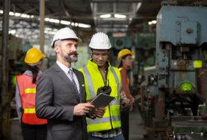 A bearded man in a suit and hard hat holds a tablet while standing in a factory, discussing employee compliance with a worker in a high-visibility vest and hard hat. Two other workers in high-vis vests and hard hats are seen in the background amid industrial machinery, ensuring safety culture is maintained.