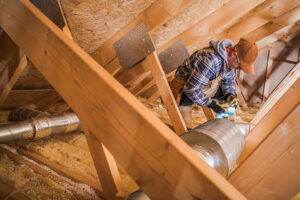 A worker wearing a checked shirt, protective glasses, gloves, and a cap is installing or inspecting silver air ducts in an attic with exposed wooden beams. Mindful of the hazards in such confined spaces, he navigates around yellow foam insulation and scattered material.