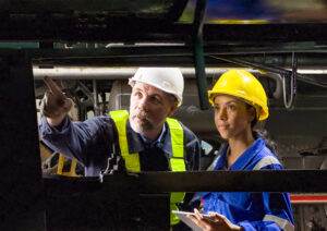 Two workers in safety helmets and vests are inspecting machinery. The man on the left is pointing at something, while the woman on the right holds a tablet. They appear to be in an industrial setting, focused on conducting a risk evaluation and discussing possible mitigation measures for the equipment.