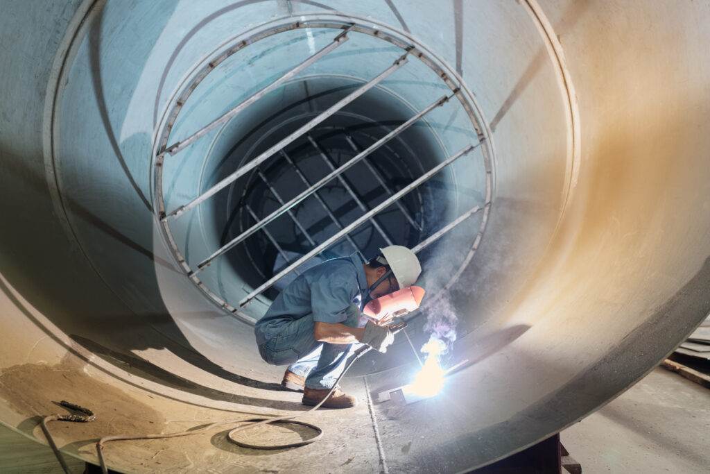 A worker in a hard hat and safety gear is welding the interior of a large metal pipe, handling the challenges of confined space work. The scene is illuminated by the bright light from the welding torch. The surrounding area appears industrial, with large metal structures.