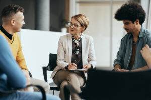 A group therapy session focused on mental health in the workplace features a middle-aged woman in a blazer leading the discussion, holding a notepad and pen. She is seated between two men, one in a yellow sweater and the other in a blue shirt. They are all seated in a circle of chairs in a well-lit room.