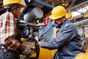 Two industrial workers wearing yellow hard hats and work gloves are operating machinery in a factory. They are focused on adjusting equipment, emphasizing proper machine guarding, and appear to be engaged in a task that involves heavy machinery and parts. The background shows additional industrial equipment.