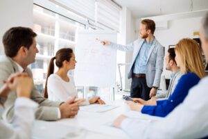 A man stands and points to a flowchart on a whiteboard during a business meeting. Four colleagues, two men and two women, are seated around a table, listening and engaging. The room has large windows with a cityscape view in the background, reflecting the organization’s strong safety culture.