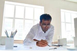 A man with glasses, dressed in a white shirt, meticulously studies and draws on architectural blueprints spread out on a bright, white desk. Several pencils and model buildings are arranged around him like a training matrix, and large windows provide natural light in the room.