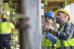 Two new construction workers wearing helmets and high-visibility vests use a spirit level to check a concrete column on a construction site. One worker in a blue helmet and glasses, another in a yellow helmet. A third worker is seen in the background working, highlighting critical safety training.
