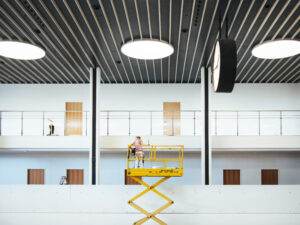 A worker in a pink shirt stands on a yellow scissor lift, performing maintenance in a spacious, modern building with high ceilings, large circular lights, and a large wall clock. This well-thought-out work environment also features fall protection systems to ensure safety. Another worker is visible in the background on the upper level.