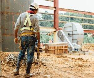 A construction worker in a safety harness and helmet stands on a muddy construction site. They are holding a tool while facing away from the camera, demonstrating proper PPE use. The site has scattered cables, a large fan-like machine, and partially constructed steel structures in the background.