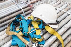 A white safety helmet and a blue and yellow safety harness, essential fall protection equipment, are placed on a pile of metal pipes at the construction site. The gear appears ready for use in ensuring worker safety through continuous inspection.