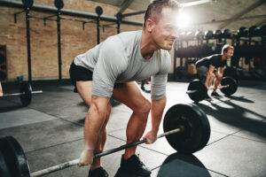 A man in a gym wearing a gray shirt and black shorts is lifting a barbell with weights, showcasing his physical fitness. In the background, another person is also doing a similar exercise. The gym has a brick wall, large windows, and various fitness equipment.