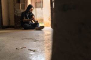 A person with long hair sits on the floor in a dimly lit, abandoned building. Two used syringes lie on the ground in the foreground. Partially blurred, they focus on their arm, highlighting the grim reality of substance abuse. The atmosphere is somber and the building appears desolate.