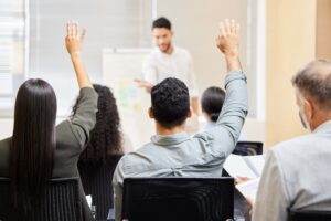 A group of people sitting in chairs faces a person standing and speaking in front of a whiteboard. Two people in the audience have their hands raised, actively engaging in employee feedback. The setting appears to be a classroom or training environment.