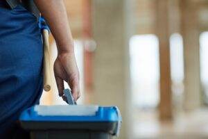 Close-up of a person in blue uniform holding a mop handle and a cleaning tool, standing next to a blue cleaning bucket with compartments, in an indoor setting with blurred background, ready to conduct their tasks effectively.