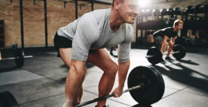 A man in a light gray shirt and black shorts lifts a barbell in a gym, showcasing his commitment to physical fitness. Others in the background engage in similar health practices. The gym, with its brick wall and racks of weights, is bathed in sunlight streaming from the left, illuminating the man’s face.