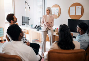 A woman in business attire stands in front of a group of four people seated in an office setting, explaining key strategies to enhance their workflow. The office features desks with computers, corkboards with papers, and a modern, minimalistic design.