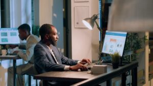 A man in a suit works at a computer on a desk in a dimly lit office. The screen displays charts and graphs related to OSHA record-keeping requirements. On the desk, there is a lamp, a potted plant, and some office supplies. In the background, another person is working at a desk, planning for 2024 compliance.