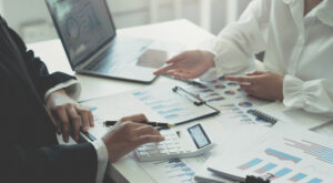 Two people at a desk are engaged in a comprehensive analysis, reviewing financial charts and graphs. One person uses a calculator while the other gestures towards the documents. A laptop displays more charts in the background, with various papers and a clipboard spread across the desk.