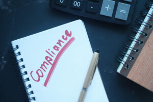 A notepad with the word "Compliance" written in red capital letters is placed on a dark surface. Next to the notepad is a beige pen, a black calculator, and another spiral-bound notebook with a wooden cover—perfect tools for ensuring adherence to OSHA regulations.