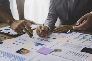 A group of people collaboratively work at a table covered with various charts, graphs, and documents. They use pens to point out and discuss different data points and trends, highlighting their focused analysis of workplace falls. Post-it notes are scattered around the papers, contributing to their data-driven approach.