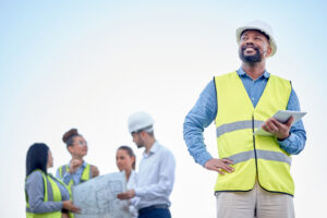 A person in a yellow safety vest and white hard hat holds a tablet and looks up while smiling, exemplifying workplace safety. In the background, four people in safety vests and hard hats discuss blueprints against a clear sky.