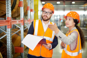 Two workers in orange safety vests and hard hats stand in a warehouse lined with shelves. One holds a document and appears to listen attentively, while the other points toward something off-camera, suggesting a discussion or inspection is taking place in this industrial setting.