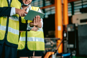 Two workers wearing yellow safety vests and helmets stand in an industrial setting, reviewing information on a tablet. One person points decisively at the screen while the other looks on attentively. With equipment and machinery in the background, their focus is on workplace safety to prevent trips, slips, and falls.