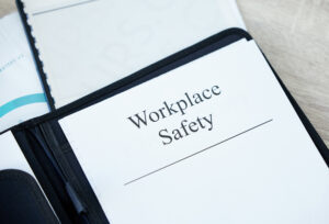 An open binder with a section titled "Workplace Safety" is displayed, highlighting upcoming OSHA changes for 2024. The binder is black, and the page is white with printed text. Other documents and papers are partially visible in the background.